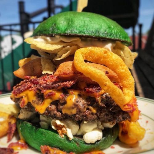 A towering burger with a green bun, loaded with cheese, bacon, onion rings, and chips, served outdoors.