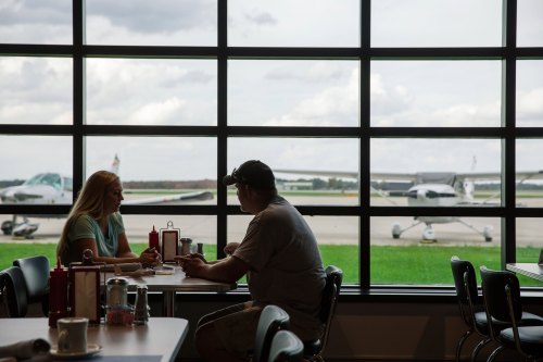 Two people sit at a table in a diner, chatting with a view of airplanes outside through large windows.