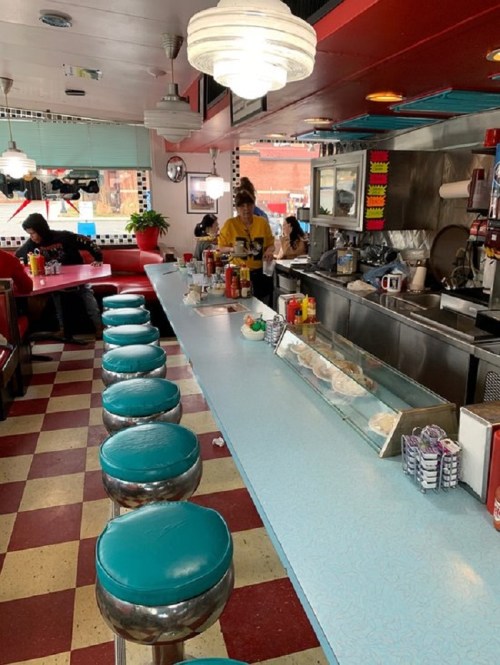 A retro diner interior with turquoise stools, a long counter, and a waitress serving customers in a cozy atmosphere.