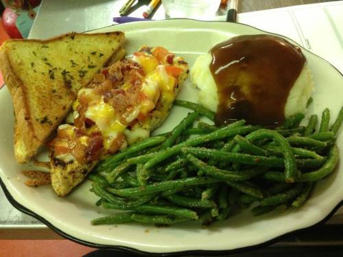 A plate of grilled chicken topped with cheese and bacon, served with green beans, mashed potatoes with gravy, and toast.