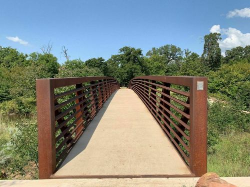 A metal bridge with a slatted railing spans a path through lush greenery under a clear blue sky.