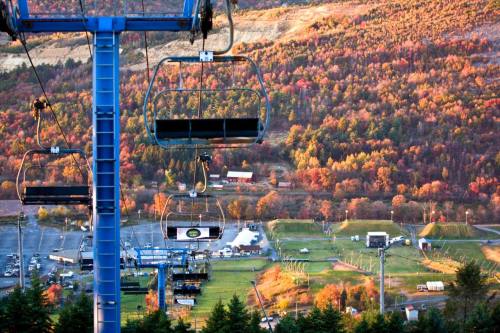 A ski lift with empty chairs overlooks a colorful autumn landscape and a parking area below.