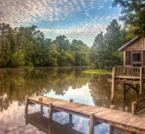 A serene river scene with a wooden dock, lush greenery, and a rustic cabin reflecting in the calm water.