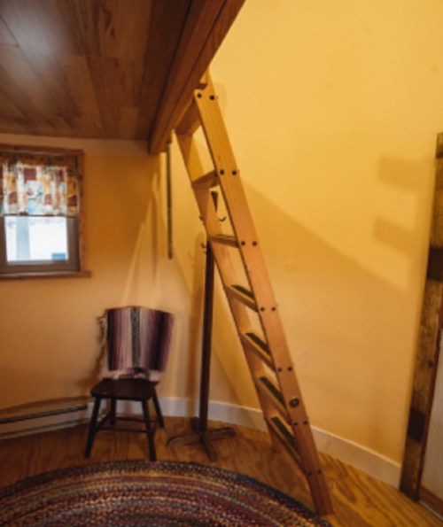 A wooden ladder leans against a wall in a cozy room with a chair and colorful rug.