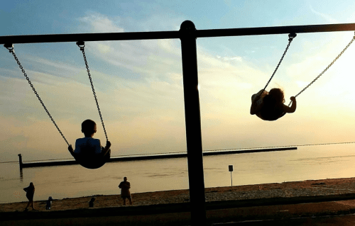 Two children swing against a sunset backdrop by the water, with silhouettes of people in the distance.
