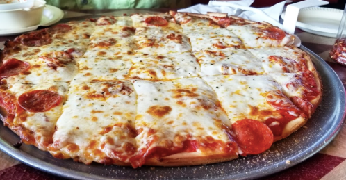 A large pepperoni pizza with melted cheese, served on a metal tray, with a red and white checkered tablecloth in the background.