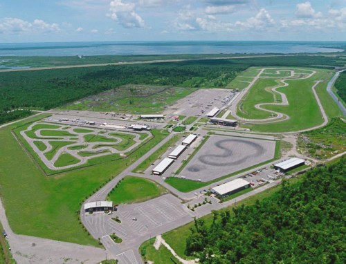 Aerial view of a large motorsport facility with multiple racetracks and parking areas, surrounded by greenery and water.