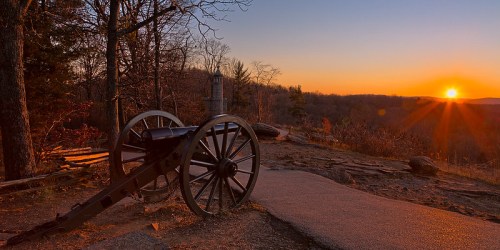 A historic cannon on a path at sunset, with trees and hills in the background, casting a warm glow.