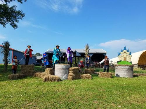 Colorful figures in costumes perform on hay bales at a farm, with a blue sky and buildings in the background.