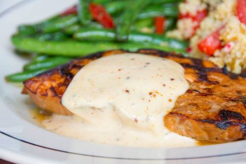Grilled salmon fillet topped with creamy sauce, served with green beans and quinoa salad on a white plate.