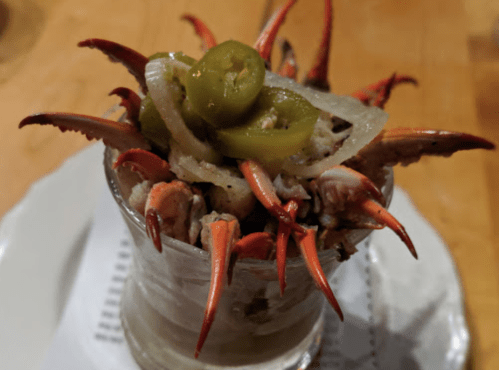 A glass dish filled with seafood, topped with jalapeños and onions, featuring crab claws protruding from the top.