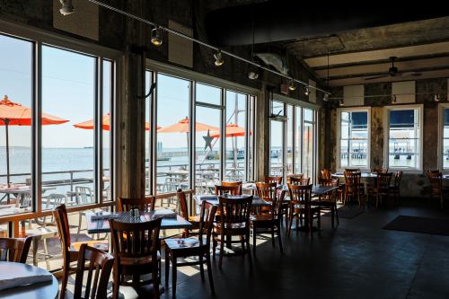Bright restaurant interior with wooden tables and chairs, large windows, and orange umbrellas overlooking a waterfront.