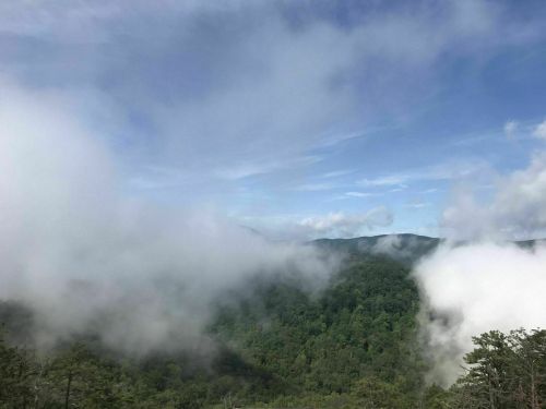 A misty landscape of green mountains under a blue sky, with clouds partially obscuring the view.