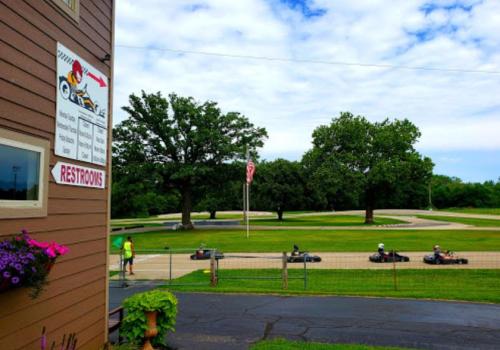 A go-kart track with racers on the course, viewed from a building with a restroom sign and greenery in the foreground.