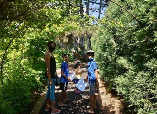 A family of four wearing masks walks through a lush, green garden path surrounded by trees and foliage.