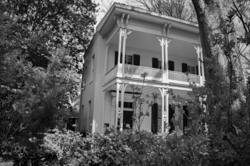 A historic two-story house with white columns, surrounded by greenery, captured in black and white.