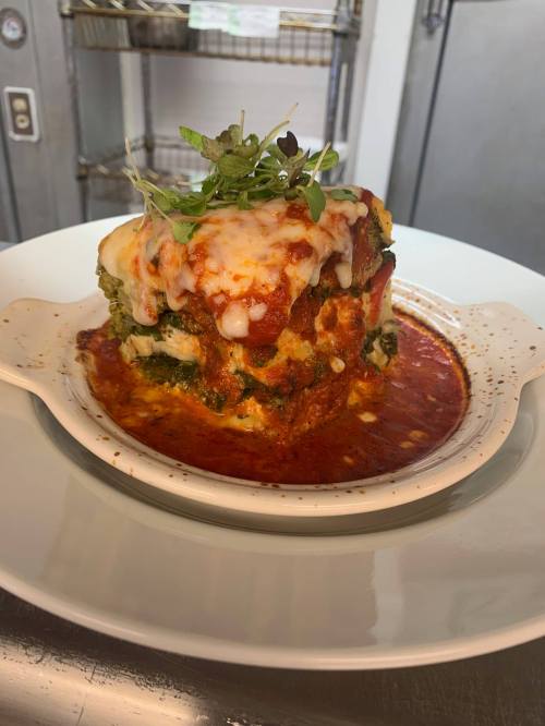 A layered vegetable lasagna topped with melted cheese and garnished with microgreens, served in a white dish.