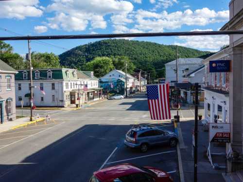 A small town street with a large American flag, shops, and a backdrop of green hills under a blue sky.