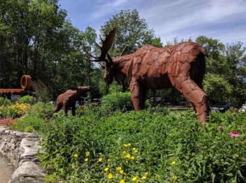 A large metal moose sculpture stands beside a smaller bear sculpture, surrounded by colorful flowers and greenery.