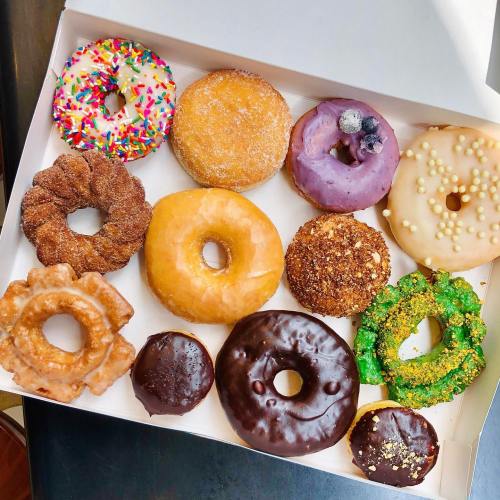 A box of assorted donuts, featuring various flavors and toppings, including sprinkles, chocolate, and fruit.