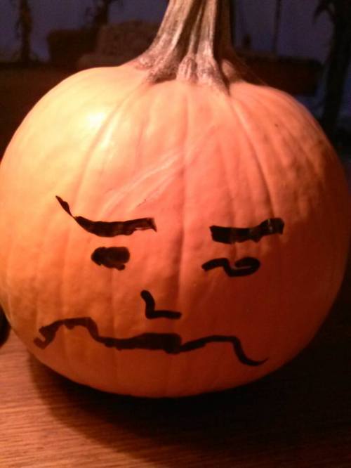 A carved pumpkin with a drawn face showing a frown, resting on a wooden surface.