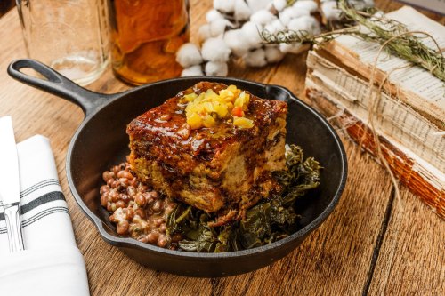 A cast iron skillet holds a slice of cornbread topped with pineapple, served on a bed of collard greens and beans.