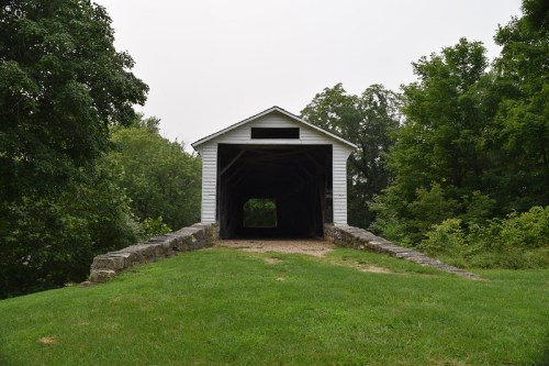 A white covered bridge with a stone foundation, surrounded by green trees and grass, leading into a dark interior.