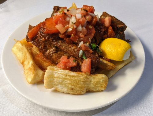 A plate of grilled meat topped with fresh salsa, served with fried plantains and a lemon wedge.