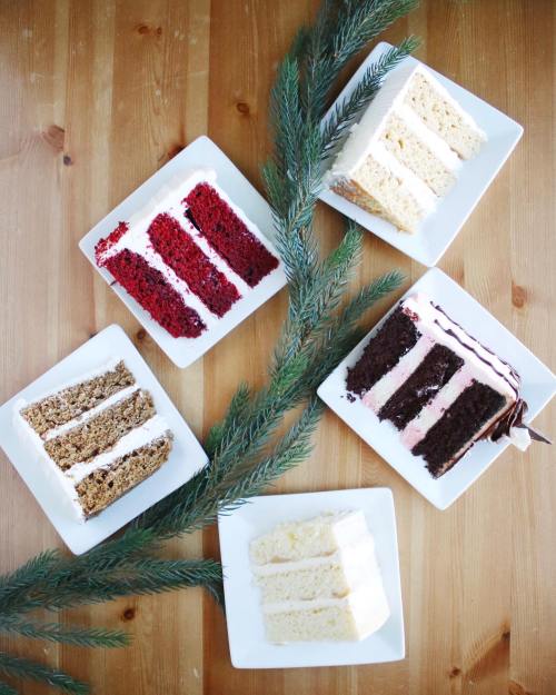 Five slices of cake on white plates, including red velvet, chocolate, and vanilla, arranged with pine branches on a wooden table.