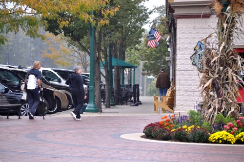 A woman walks with a walker, while another person strolls nearby in a quaint outdoor shopping area with flowers.