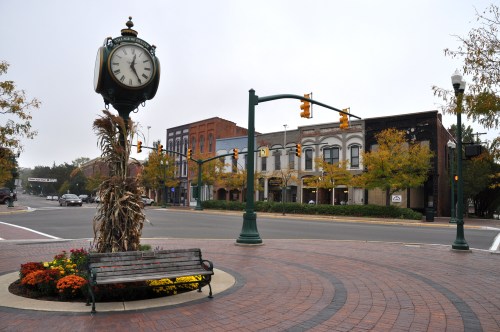 A vintage clock stands in a roundabout surrounded by colorful flowers and trees, with shops lining the street in the background.