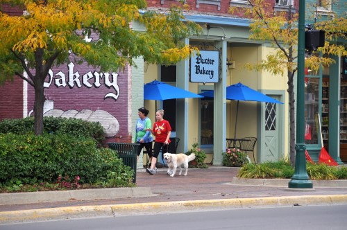 Two women walk a dog past a bakery with blue umbrellas, surrounded by autumn foliage in a charming town setting.