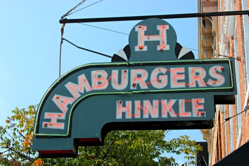 Vintage neon sign for "Hinkle Hamburgers" against a clear blue sky, surrounded by green foliage.