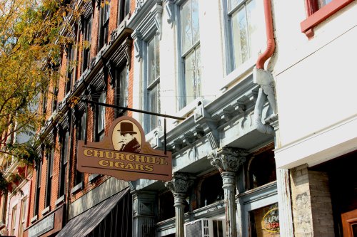 Sign for "Churchill Cigars" hanging from a historic building with ornate architecture and autumn foliage nearby.