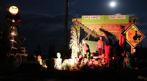 A nighttime scene of a harvest display with pumpkins, scarecrows, and a sign reading "Eat Locals" under a full moon.