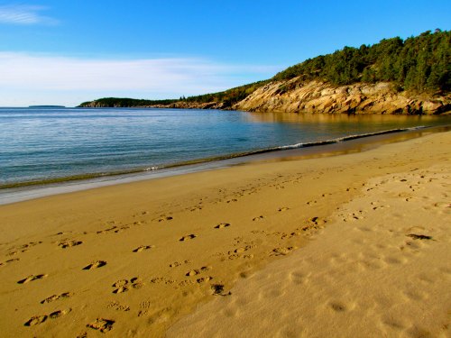 A serene beach scene with footprints in the sand, calm water, and rocky hills under a clear blue sky.