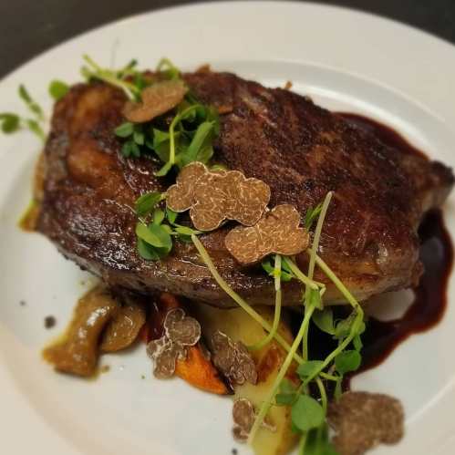 A beautifully plated steak topped with microgreens and truffle slices, served with sautéed mushrooms and a rich sauce.