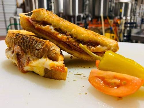 A close-up of a grilled sandwich with layers of meat and cheese, served with a slice of tomato and a pepper on the side.