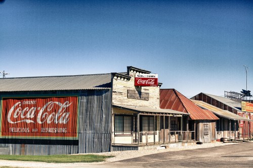 A vintage storefront with a large Coca-Cola advertisement, featuring rustic buildings and a clear blue sky.