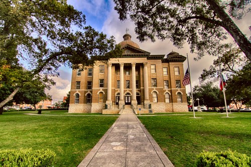Historic courthouse with a dome, flanked by trees and flags, set on a grassy lawn under a cloudy sky.