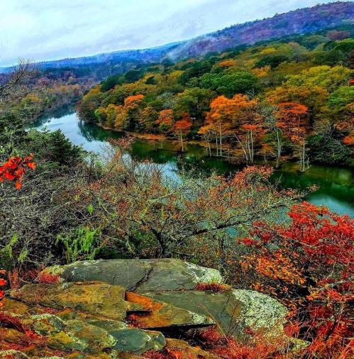 A scenic view of a river surrounded by autumn foliage and rolling hills under a cloudy sky.