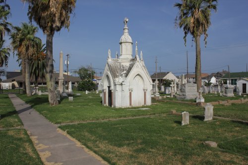 A cemetery scene featuring a decorative mausoleum surrounded by palm trees and grassy pathways.
