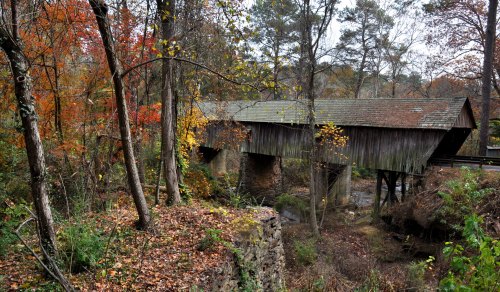 A rustic covered bridge surrounded by autumn foliage and trees, spanning a small stream in a serene landscape.