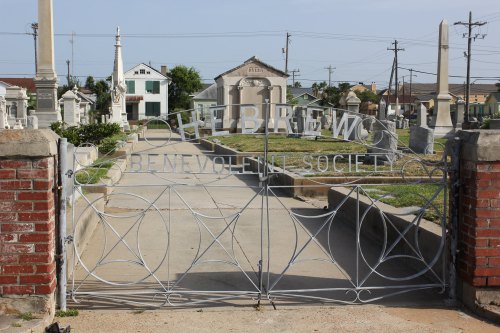 A decorative metal gate with "Hebrew Benevolent Society" inscribed, leading into a cemetery with tombstones visible.