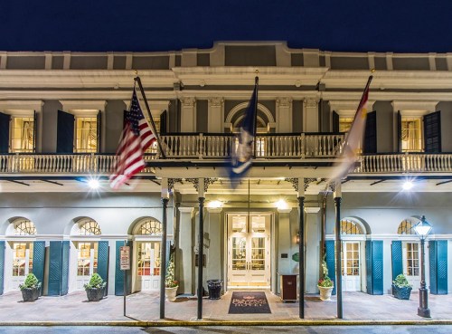 Historic hotel facade at night, featuring flags and illuminated entrance with potted plants.