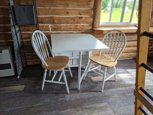 A small white table with two wooden chairs in a cozy log cabin setting. Natural light streams through a window.