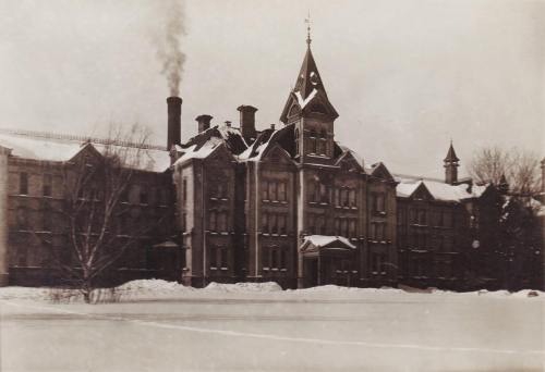 Historic building covered in snow, featuring a tall clock tower and smoke rising from a chimney. Winter scene.