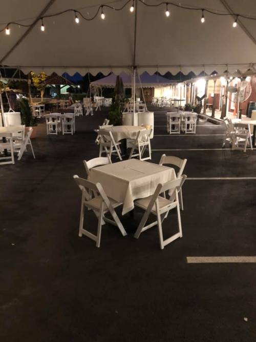 Empty outdoor dining area under a tent, featuring white tables and chairs, illuminated by string lights.