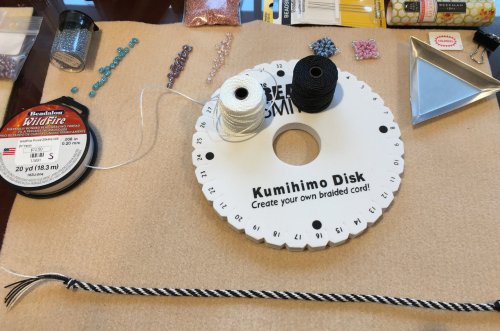 A kumihimo disk with threads, beads, and tools for making braided cord, arranged on a crafting surface.