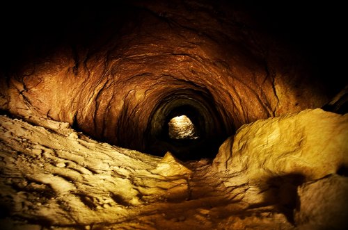 A dimly lit cave tunnel with textured walls and a distant light at the end, creating a mysterious atmosphere.